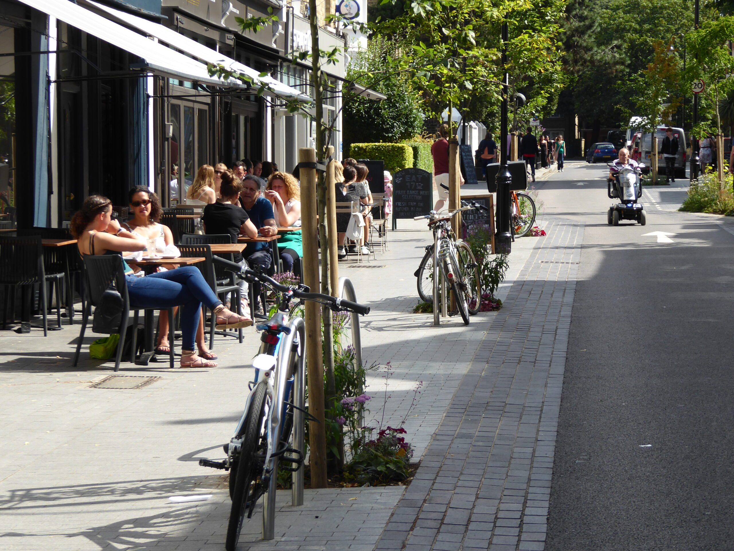 people sitting outside shops and cafes on a stepfree, broad footpath urban street with bikes parked